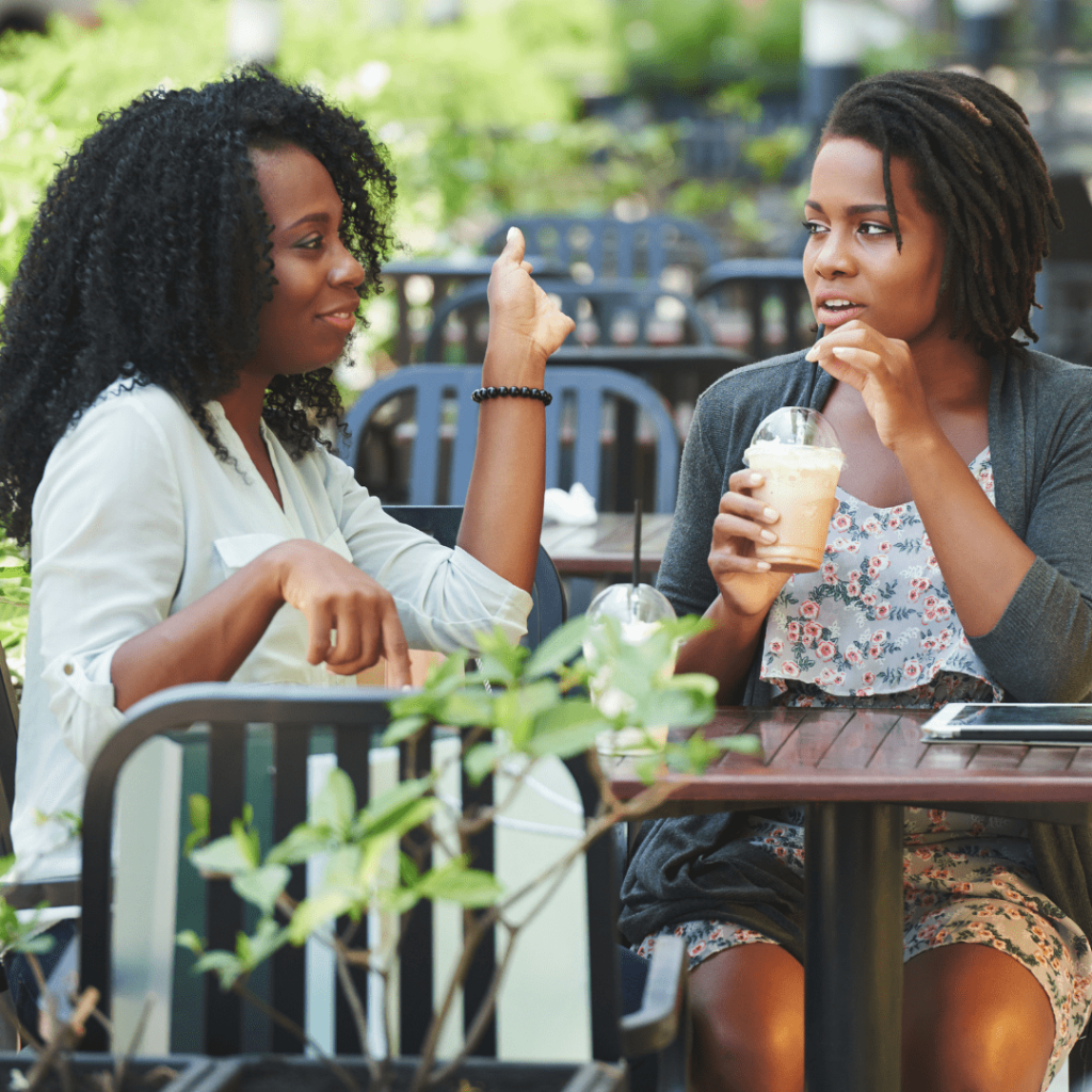 two friends sitting at an outdoor cafe talking, eating, and drinking